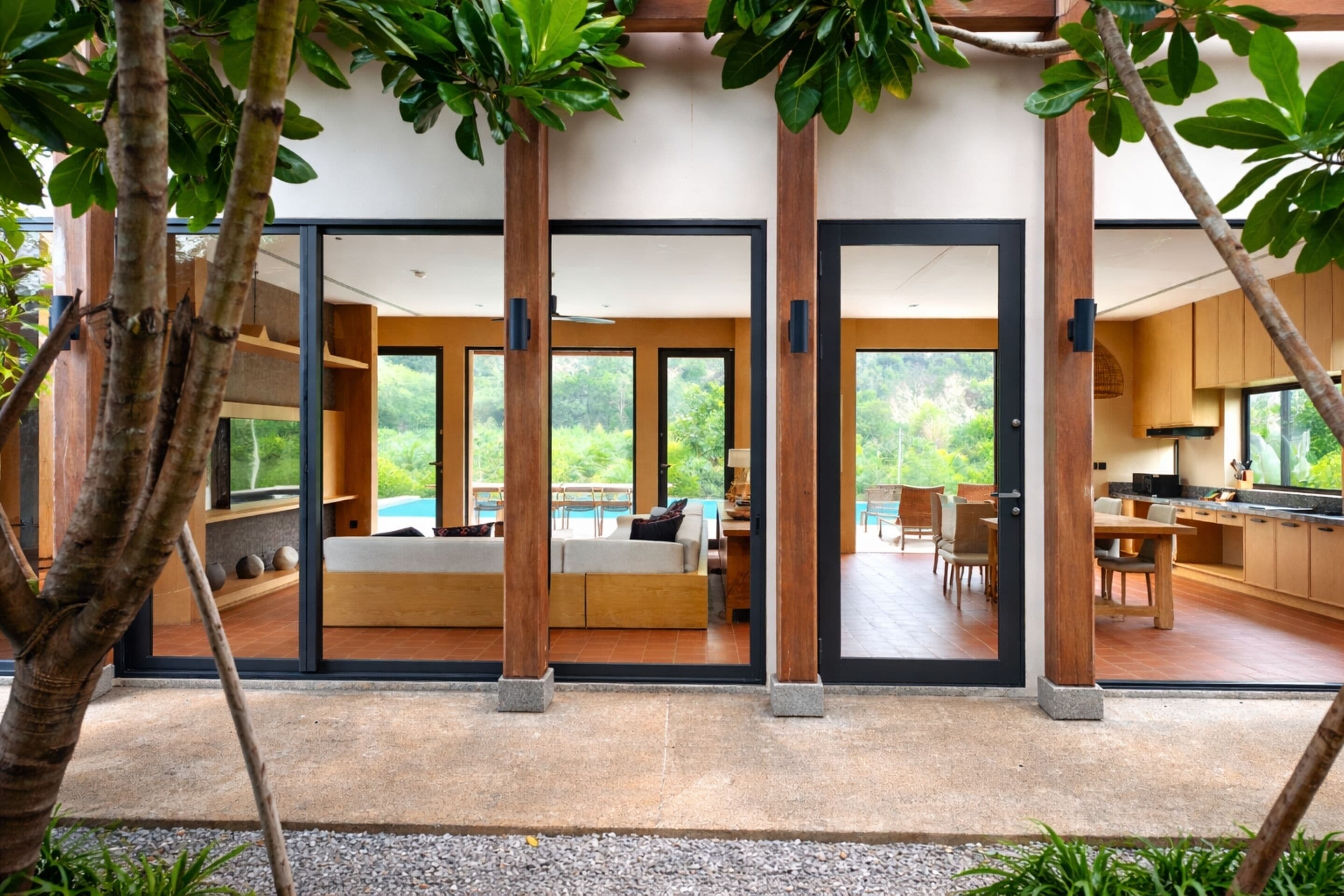 Un interior moderno con un gran ventanal corredero que da a un balcón, una zona de estar con sofá y una mesa de comedor, y una cocina visible a través de las puertas de cristal.
