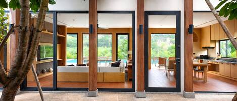 Un interior moderno con un gran ventanal corredero que da a un balcón, una zona de estar con sofá y una mesa de comedor, y una cocina visible a través de las puertas de cristal.