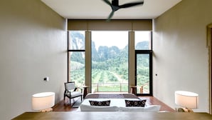 Une chambre à coucher avec un grand lit, deux tables de chevet, une chaise et une vue sur un paysage montagneux grâce à de grandes fenêtres.