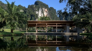 Un pavillon doté d’un toit en bois et ouvert sur les côtés, entouré d’un étang paisible dans lequel se reflètent la structure et les arbres environnants.