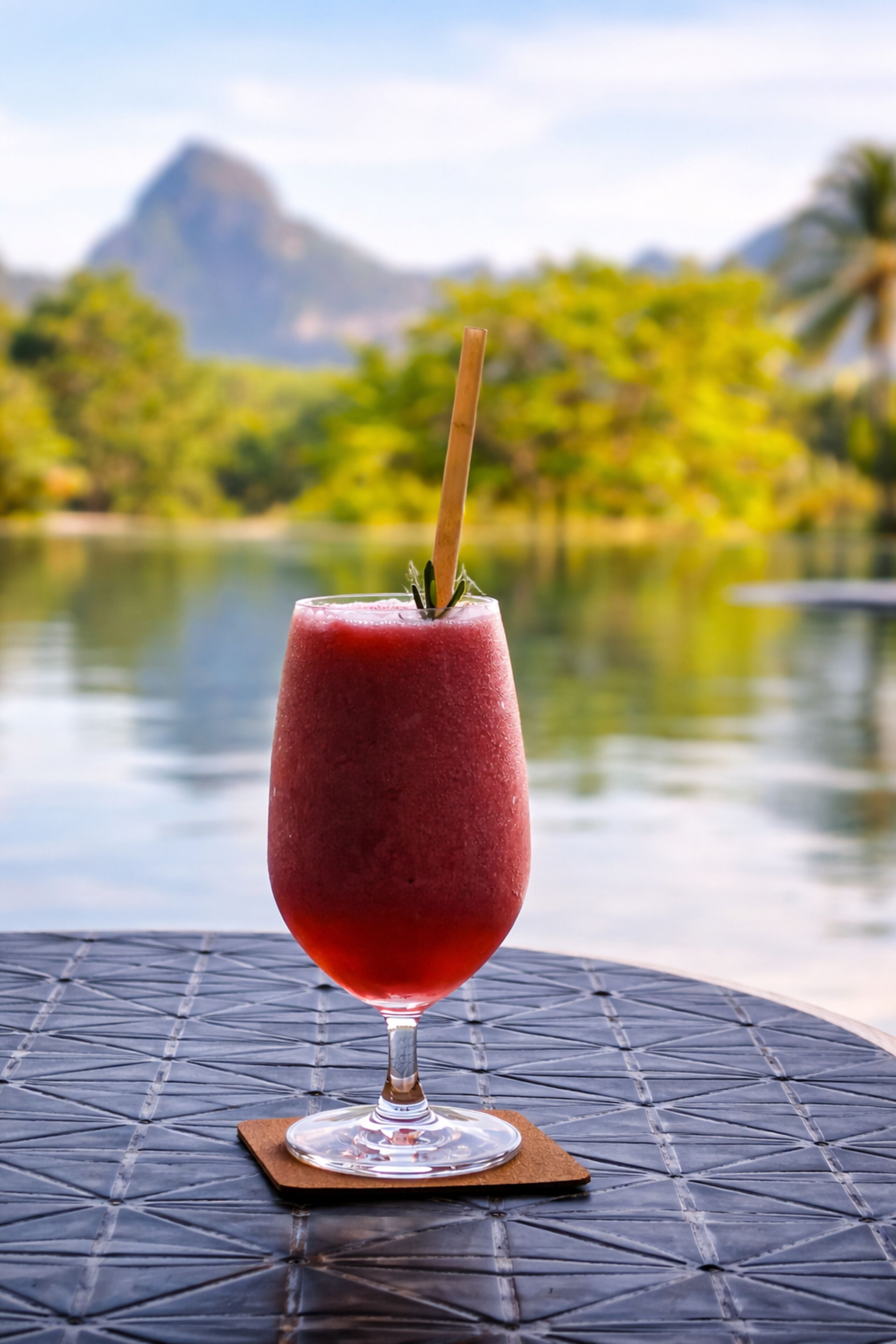 A glass of red beverage on a coaster with a mountain and greenery in the background.