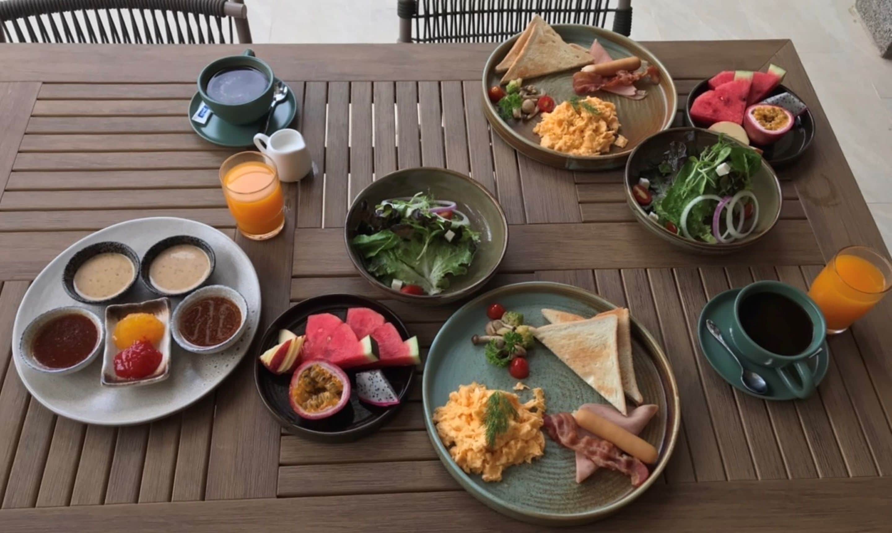 A wooden table set with breakfast items including scrambled eggs, toast, ham, and a salad.