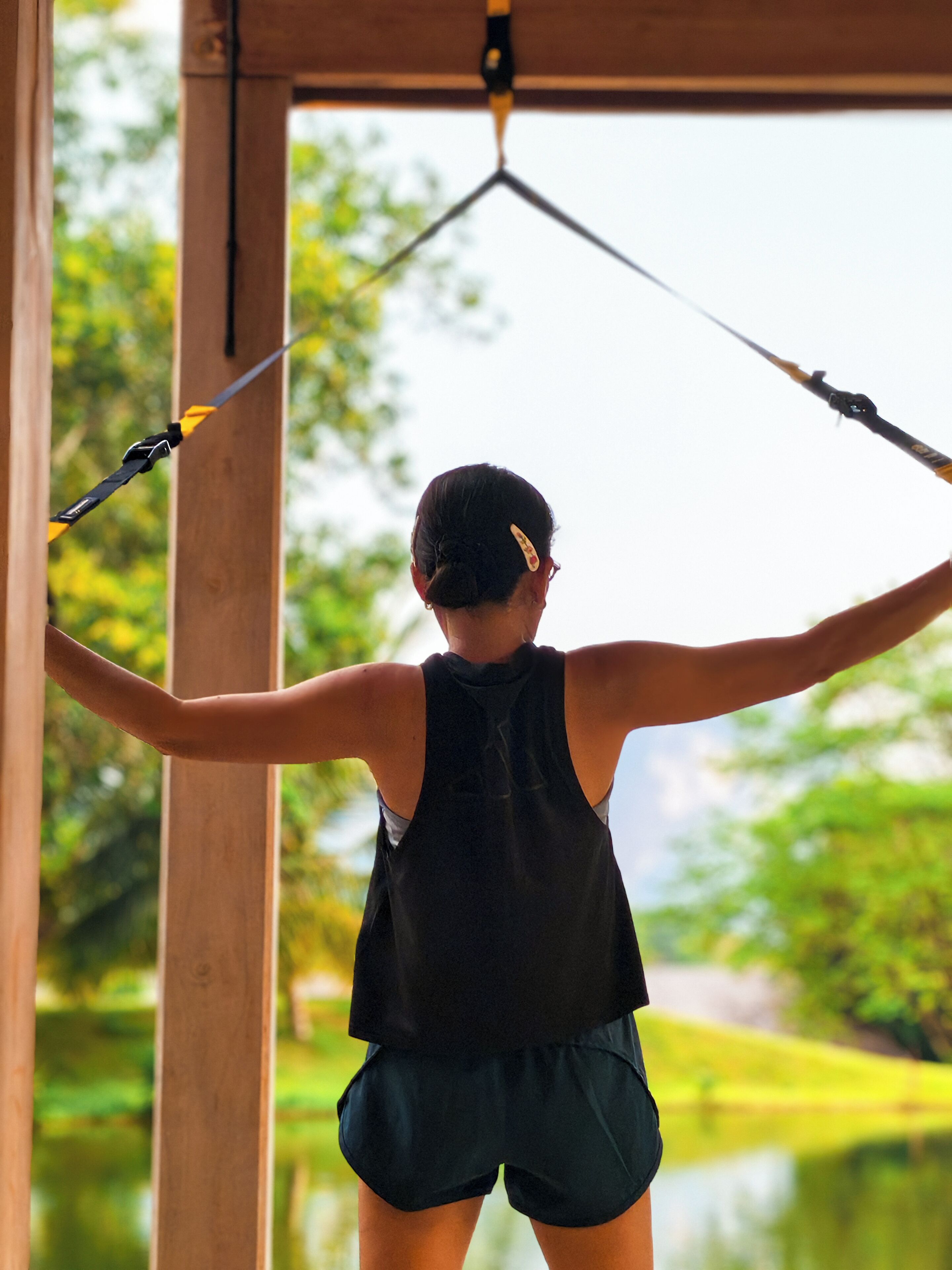 A person using a suspension training system in an outdoor setting with wooden structures and greenery in the background.