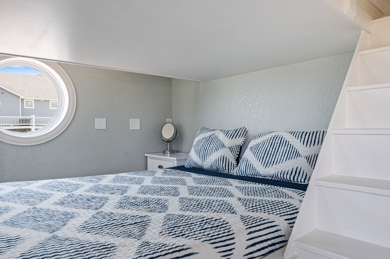 Un dormitorio con cama, una mesita de noche con espejo y una ventana con vista a una casa.