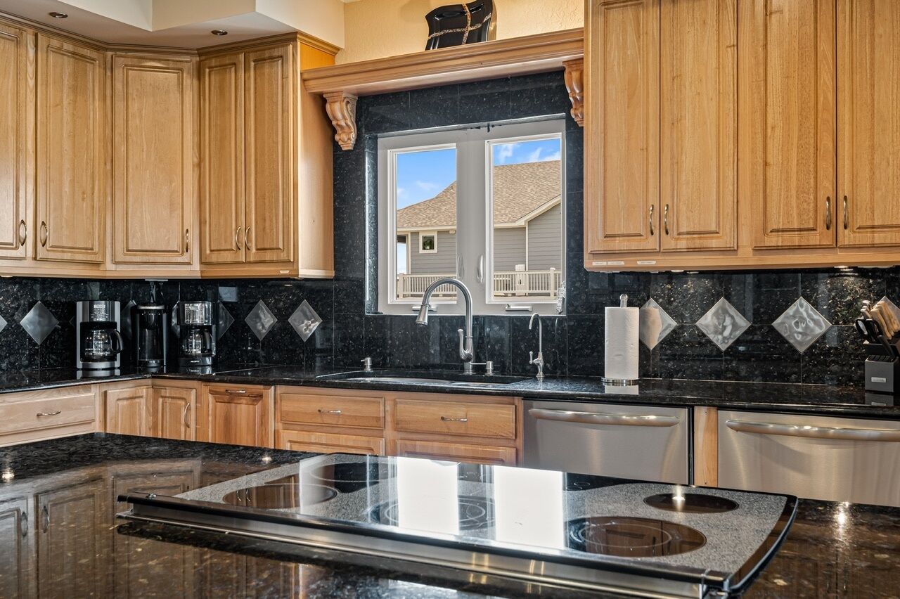 A kitchen with wooden cabinets, black granite countertops, a stainless steel stove, and a window with a view of a house.