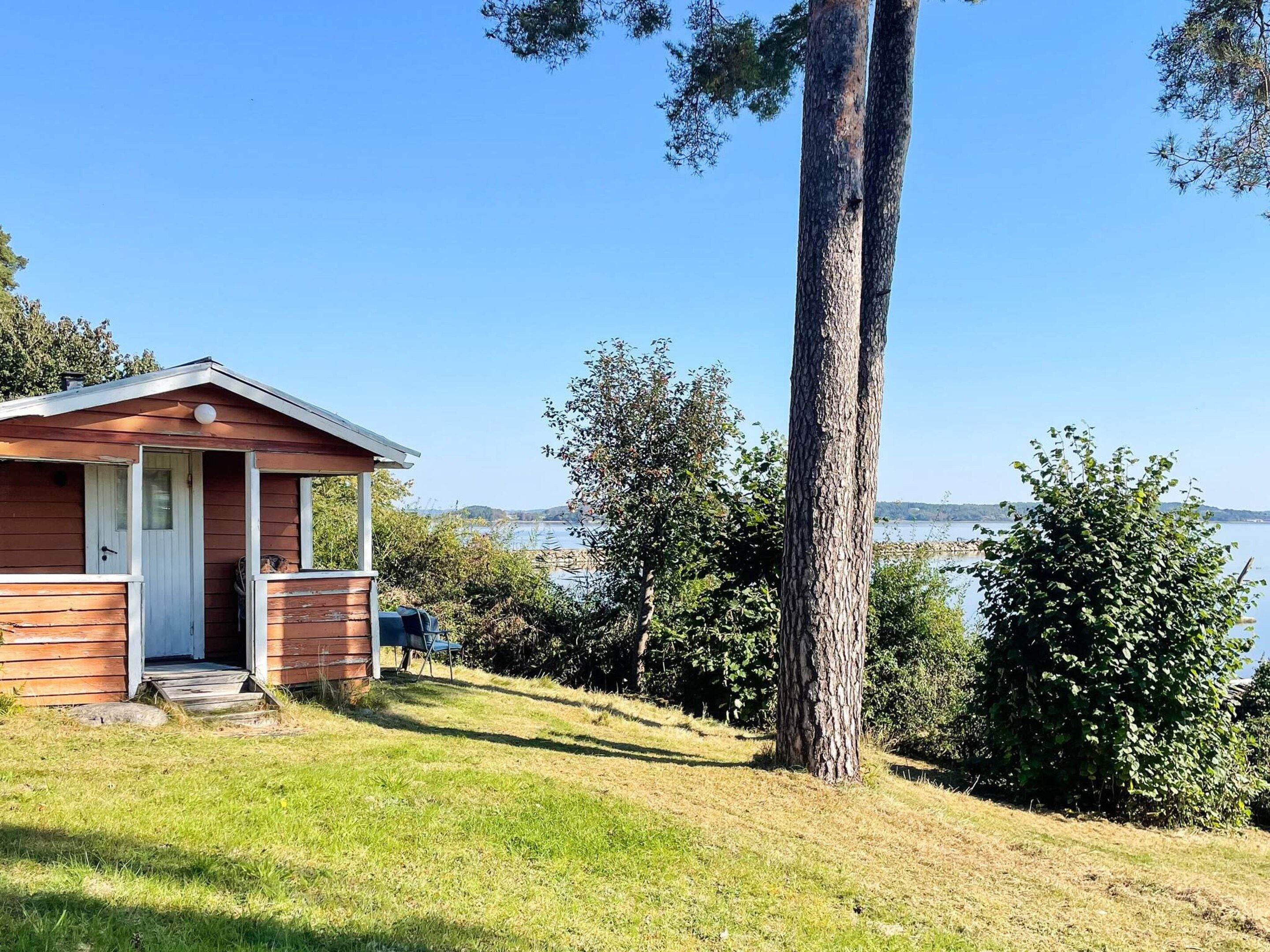 A small wooden cabin with a porch, situated on a grassy slope overlooking a body of water.