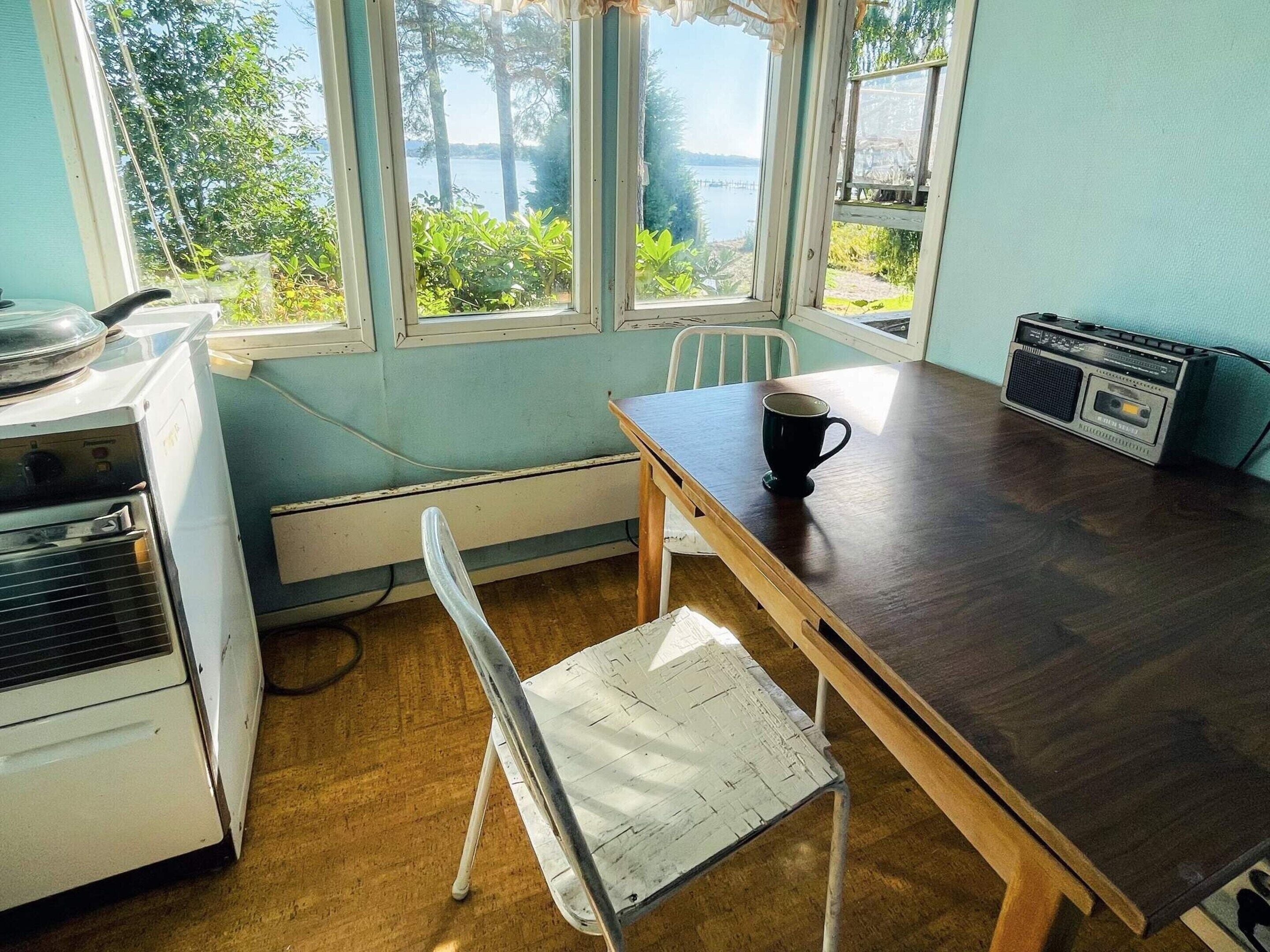 A room with a wooden table, a white chair, a radio, and a view of a lake through large windows.
