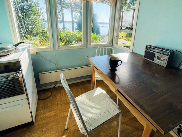 A room with a wooden table, a white chair, a radio, and a view of a lake through large windows.