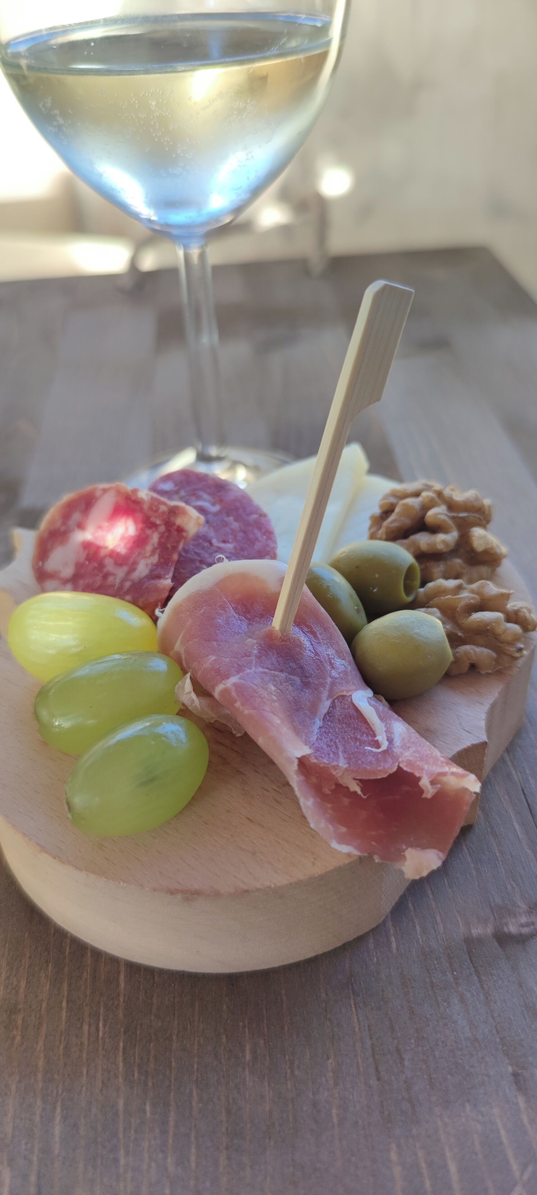 Une planche en bois sur laquelle sont disposés divers aliments, notamment des raisins, des olives, des noix et un morceau de viande, accompagnée d’un verre de vin blanc.