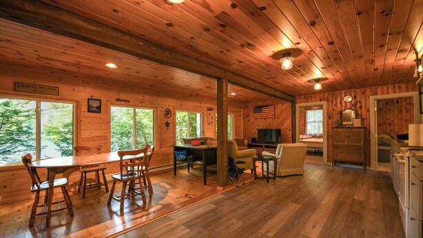 A cabin interior with wooden walls and floors, a dining area with a table and chairs, a living area with a sofa and TV, and a kitchen with cabinets and appliances. - Pet-Friendly Classic Maine Camp on Quiet Coffee Pond (Casco)