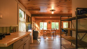 A wooden interior with a kitchen area, dining table, and chairs. Shelves and a trash bin are visible. - Pet-Friendly Classic Maine Camp on Quiet Coffee Pond (Casco)