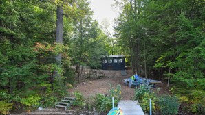 A cabin surrounded by dense forest, a dock with kayaks, and outdoor seating. - Pet-Friendly Classic Maine Camp on Quiet Coffee Pond (Casco)