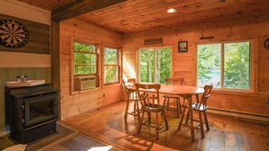 A wooden cabin interior with a dining table and chairs, a dartboard, and a wood stove. - Pet-Friendly Classic Maine Camp on Quiet Coffee Pond (Casco)