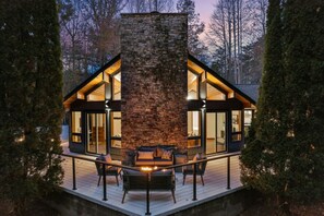 A modern cabin with a stone chimney, large windows, and a deck with outdoor seating.