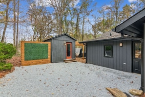 A small, dark wooden cabin with a red door, surrounded by a gravel driveway and a wooden fence with a green artificial turf section.