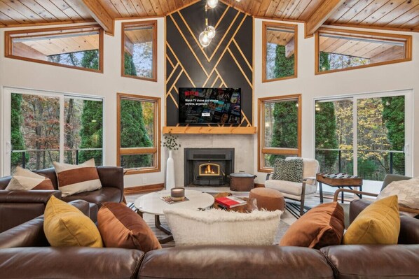 A spacious living room with a fireplace, leather sofas, and large windows offering a view of the forest.
