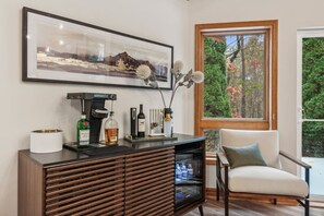 A modern living room with a large window, a framed landscape painting, a coffee machine, and a cabinet with bottles.