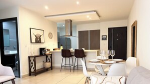 A modern living room with a sofa, a coffee table, and a dining area with a table set for two. There is a kitchen in the background with a refrigerator and a microwave. - Prime Location 2 BDR Apt, Pool, GYM, ground floor, Walkable to Everything (Santa Ana)