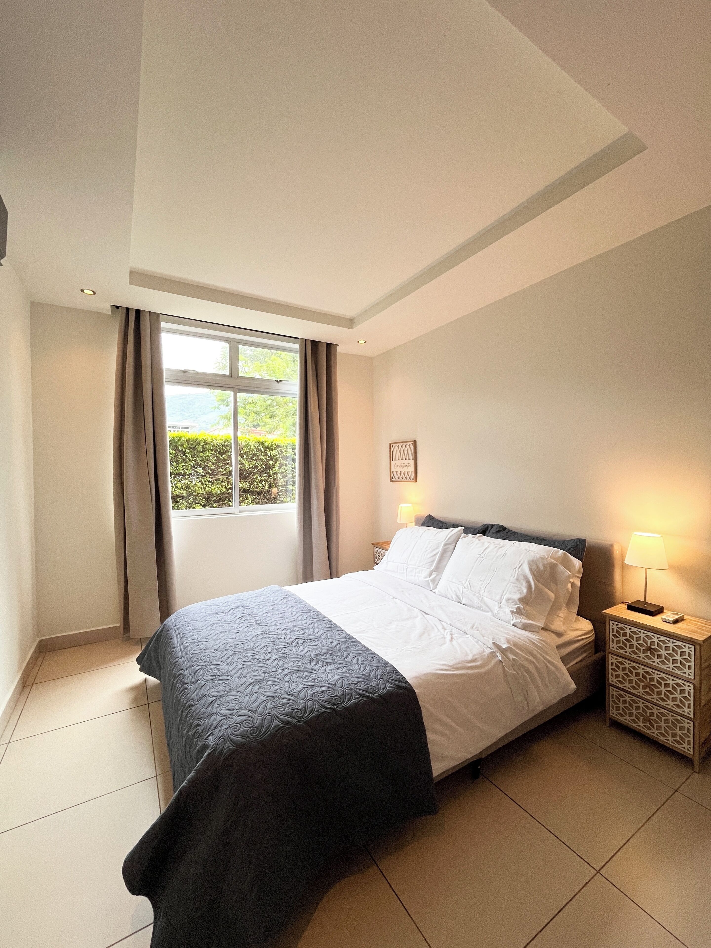 A bedroom with a bed, a nightstand, a window with curtains, and a wall-mounted picture.
