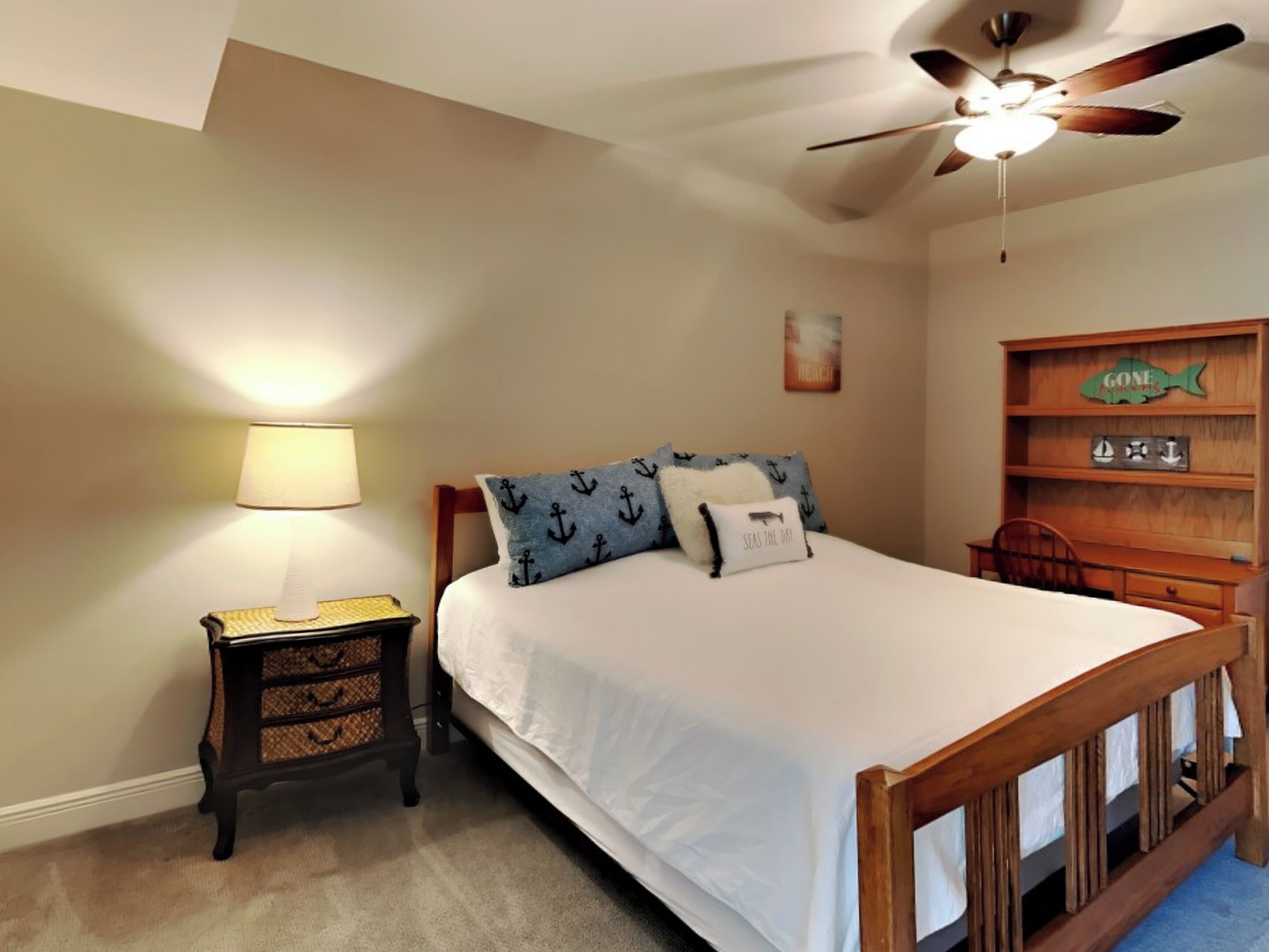 A bedroom with a bed, a nightstand, a lamp, a ceiling fan, and a wooden shelf with nautical decorations.