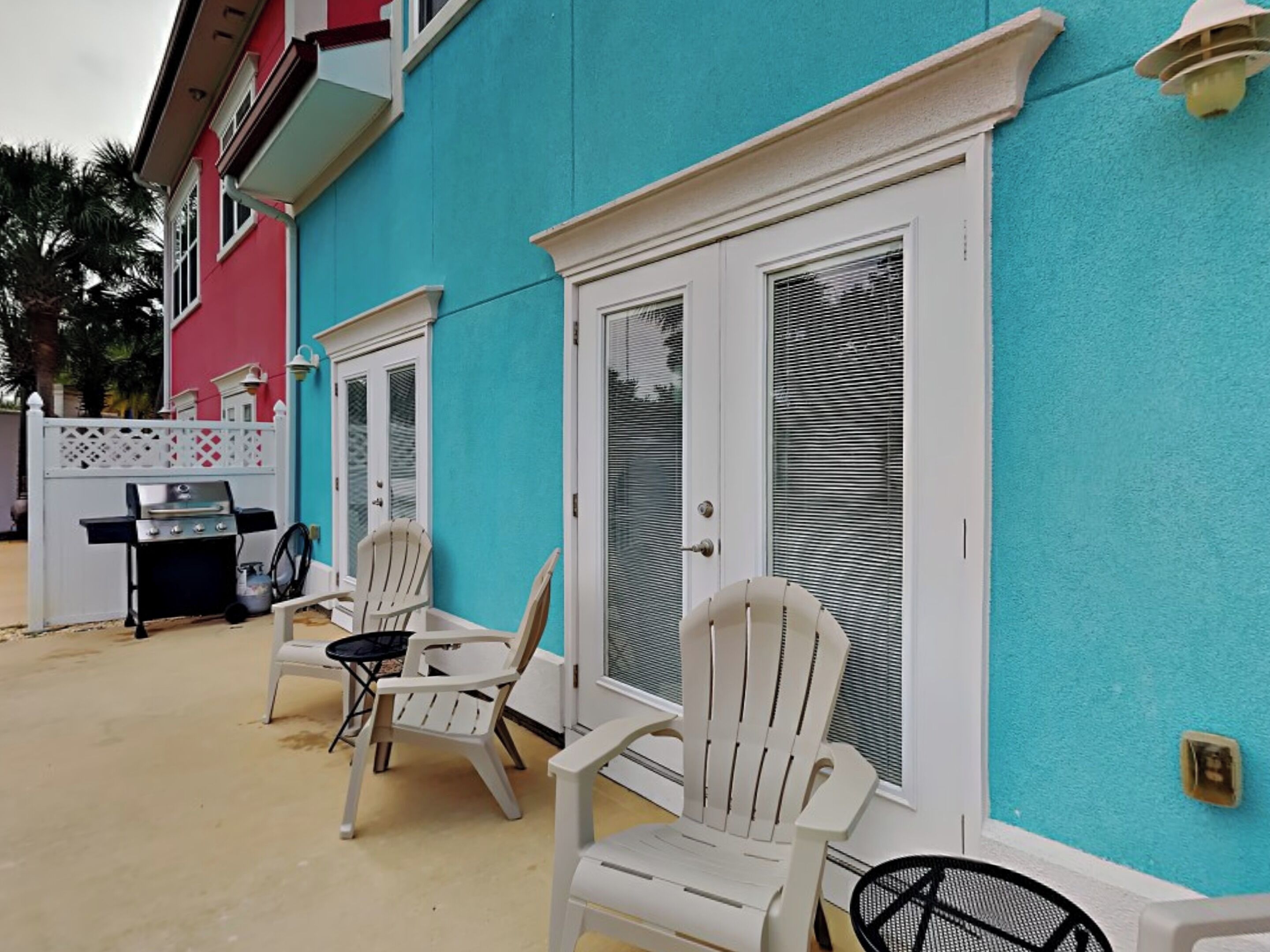 A turquoise building with white doors and windows, outdoor seating with white chairs, and a barbecue grill.