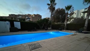 En swimmingpool med fliseterrasse, omgivet af palmer og en hæk.