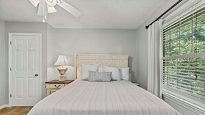 Ein Schlafzimmer mit Bett, Nachttisch, Deckenventilator, Fenster mit Jalousien und einer Tür.