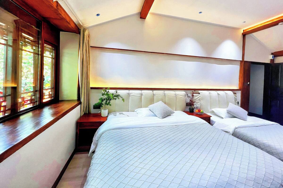 A hotel room with two beds, a large window with blinds, a bedside table with a plant, and a wooden ceiling.