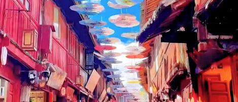 A narrow street with buildings on either side, featuring traditional architecture and multiple hanging umbrellas.