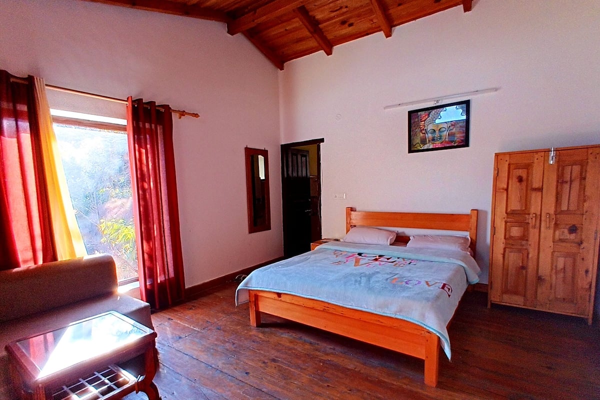 A bedroom with a wooden bed, a wooden wardrobe, a small table with a glass top, and a window with curtains.