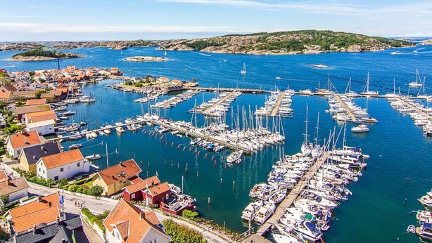 En småbåtshamn med många förtöjda båtar, omgiven av bostadshus och ett kuperat landskap i bakgrunden.