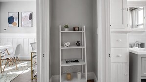 Ein helles Interieur mit einem weißen Regalsystem, einem Essbereich mit weißen Stühlen und einem weißen Badezimmer mit Waschbecken und Spiegel.