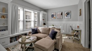 Ein Wohnzimmer mit einem beige gefärbten Sofa, einem runden Couchtisch und einem weißen Beistelltisch. Es gibt Bücherregale, ein Fenster mit Jalousien und eine weiße Heizung.