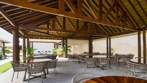 Un área de comedor al aire libre y techada con varias mesas redondas y sillas de mimbre, que cuenta con un techo de madera y plantas en macetas.