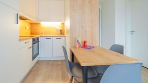 Une cuisine de taille réduite avec des meubles blancs, un plan de travail en bois et un évier. On y trouve une table à manger avec deux chaises et quelques objets posés dessus.