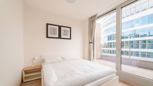Une chambre moderne avec un grand lit, une table de chevet avec une lampe et une fenêtre donnant sur un immeuble.