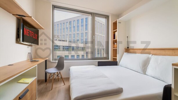 A modern hotel room with a large bed, a desk with a laptop, a chair, a TV mounted on the wall, and a window with a city view. - MyMozart City Center Residence: 702 (Prague)