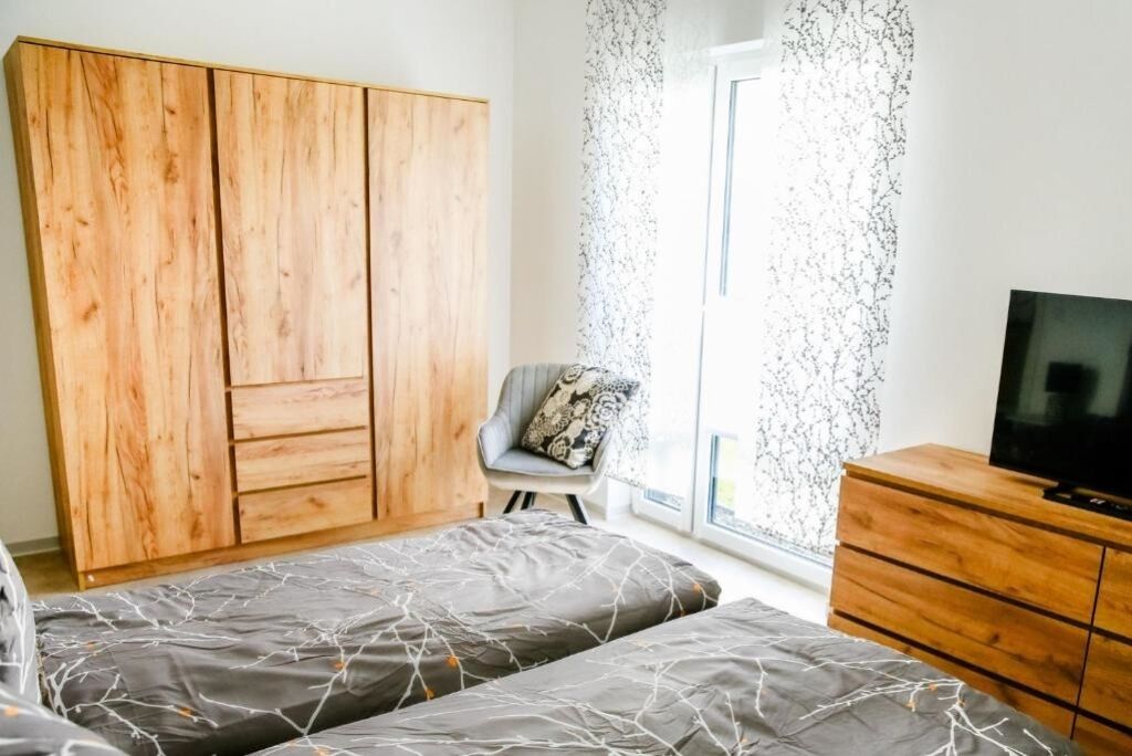 Ein Schlafzimmer mit zwei Betten, einem Holzschrank, einem Stuhl mit gemustertem Kissen, einem Fernseher auf einer Holzkommode und einem Fenster mit einem gemusterten Vorhang.