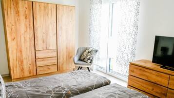Ein Schlafzimmer mit zwei Betten, einem Holzschrank, einem Stuhl mit gemustertem Kissen, einem Fernseher auf einer Holzkommode und einem Fenster mit einem gemusterten Vorhang.