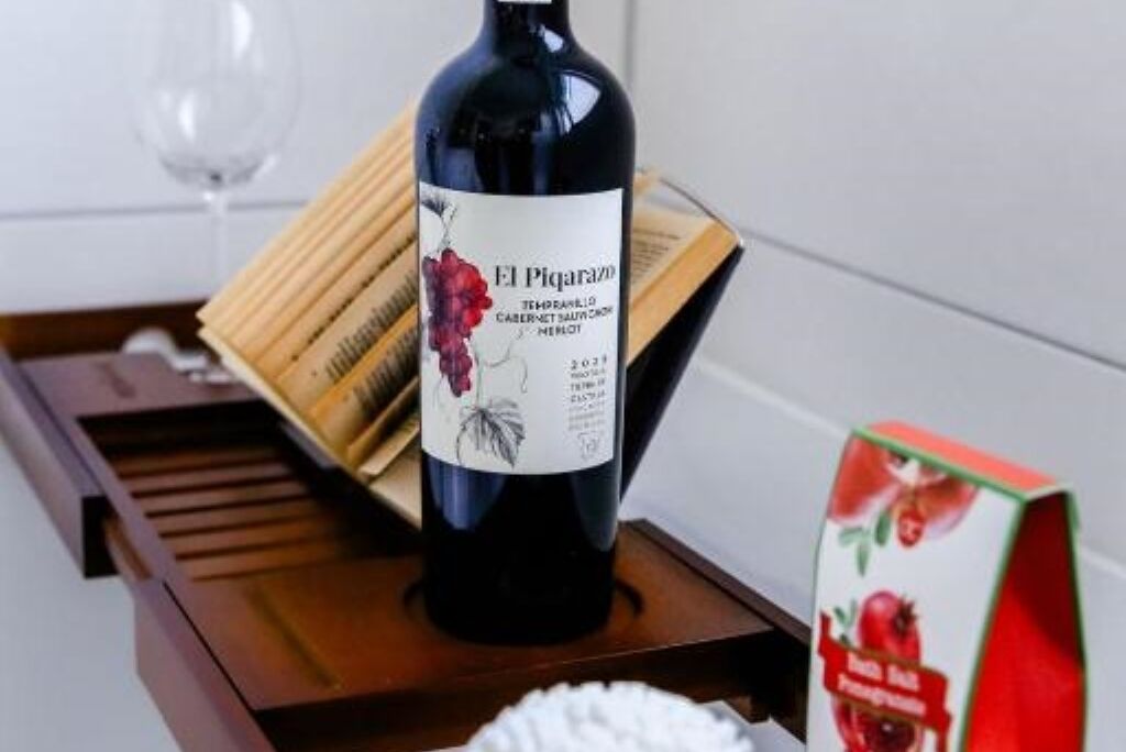 Una botella de vino El Picarazo sobre una bandeja de madera, junto con una copa de vino y una caja de zumo de granada.
