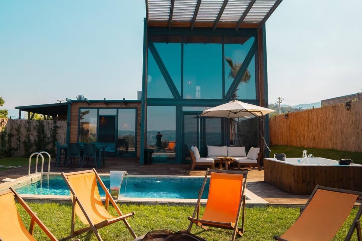 Un espace piscine extérieur moderne, doté de chaises longues, d’un jacuzzi et d’une structure fermée par des parois de verre.