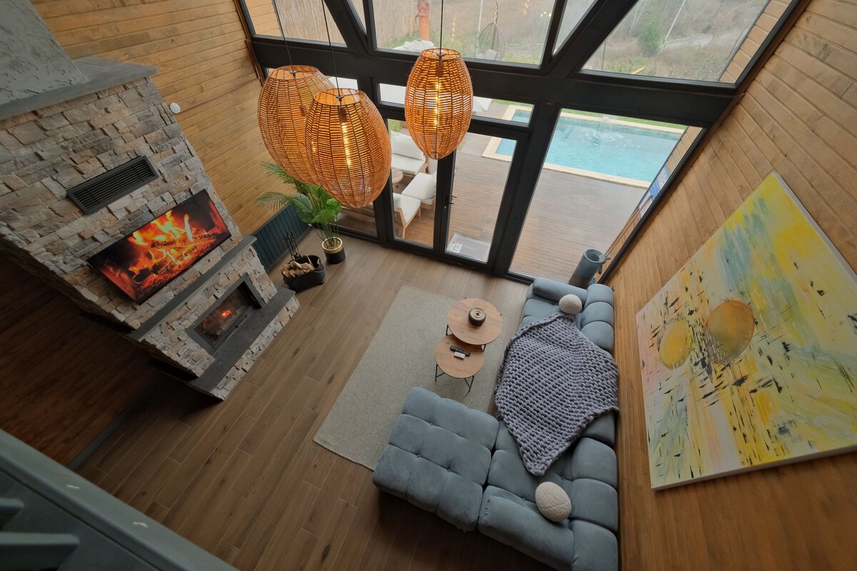 Uma sala de estar moderna com lareira de pedra, sofá cinza, mesa de centro e uma grande janela com vista para uma piscina.