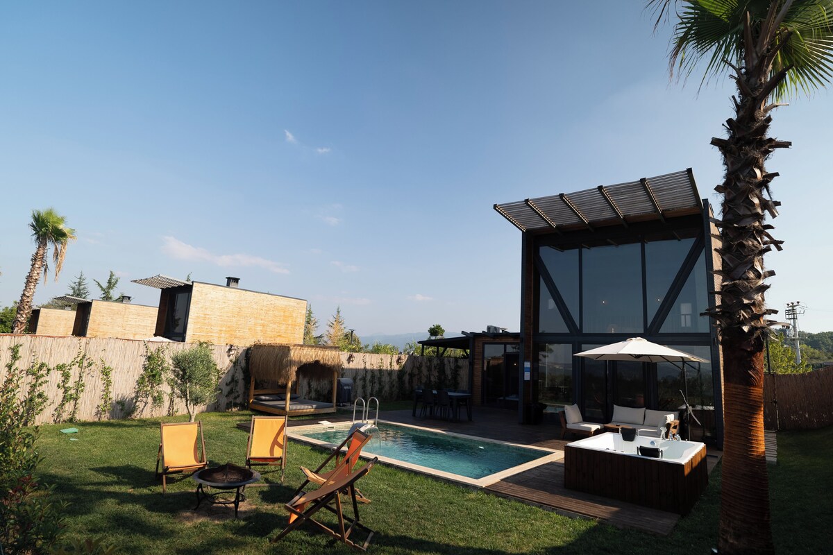 A modern outdoor pool area with a glass-enclosed lounge, seating, and a fire pit.