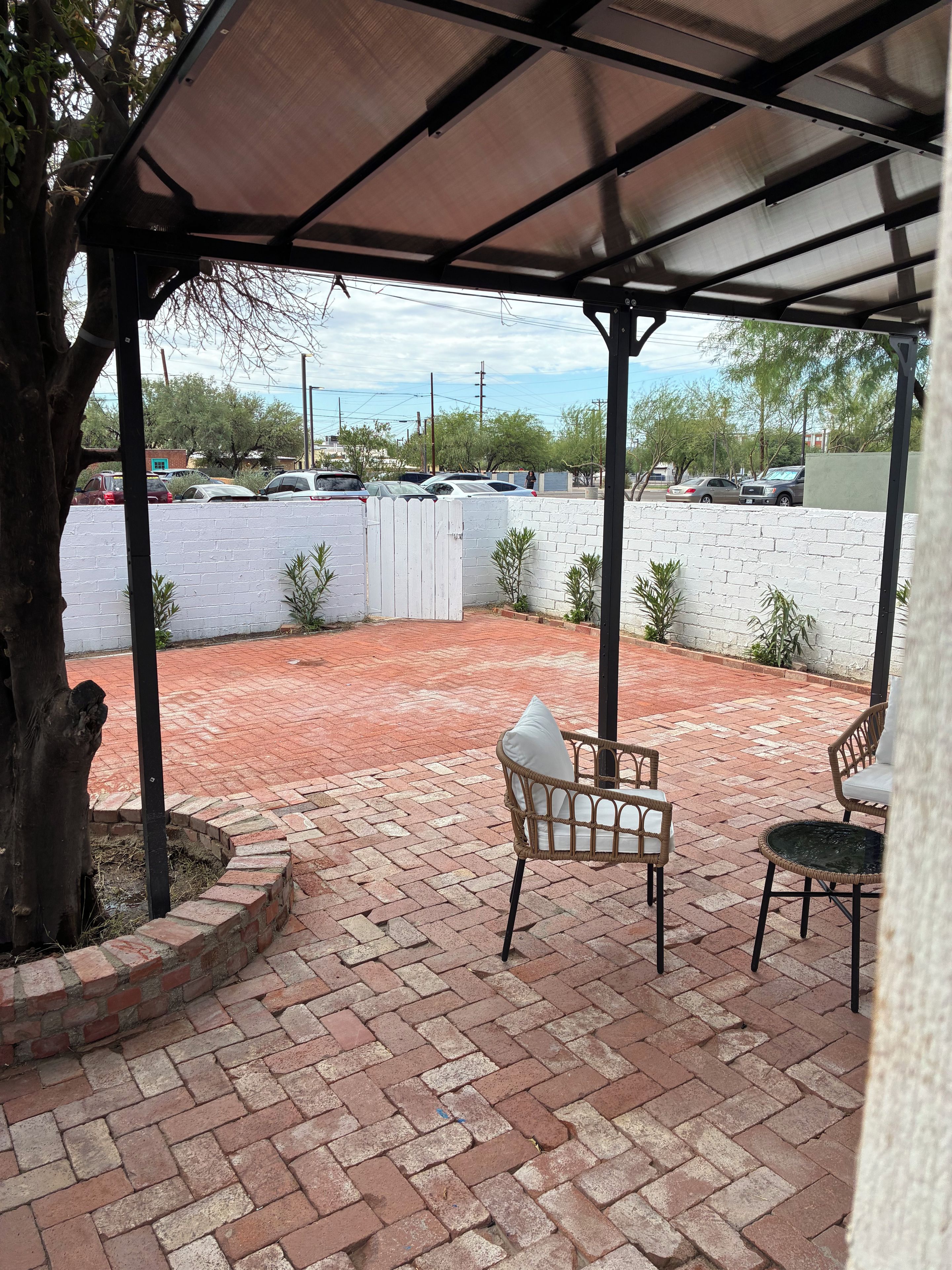 A covered patio area with brick flooring, wicker furniture, and a view of a parking lot.