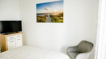 A bedroom with a bed, a television on a wooden cabinet, a grey upholstered chair, and a wall-mounted picture of a coastal landscape.