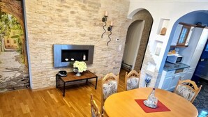 Ein Essbereich mit einem runden Holztisch, vier Stühlen, einem kleinen Beistelltisch mit Blumenvase und einer Steinwand, an der ein Fernseher hängt.