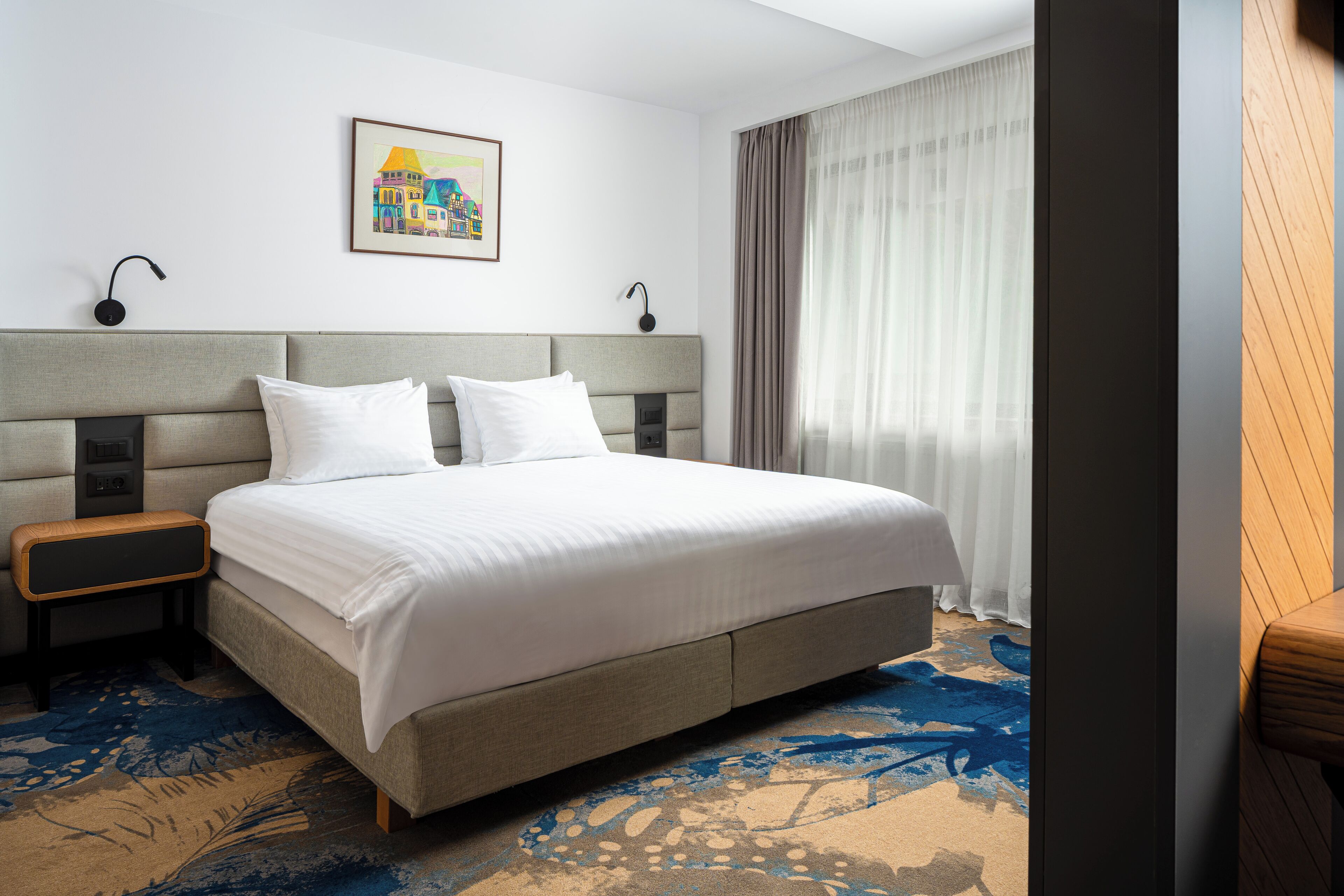 Une chambre d’hôtel avec un grand lit, une table de chevet, une lampe fixée au mur et un tableau.