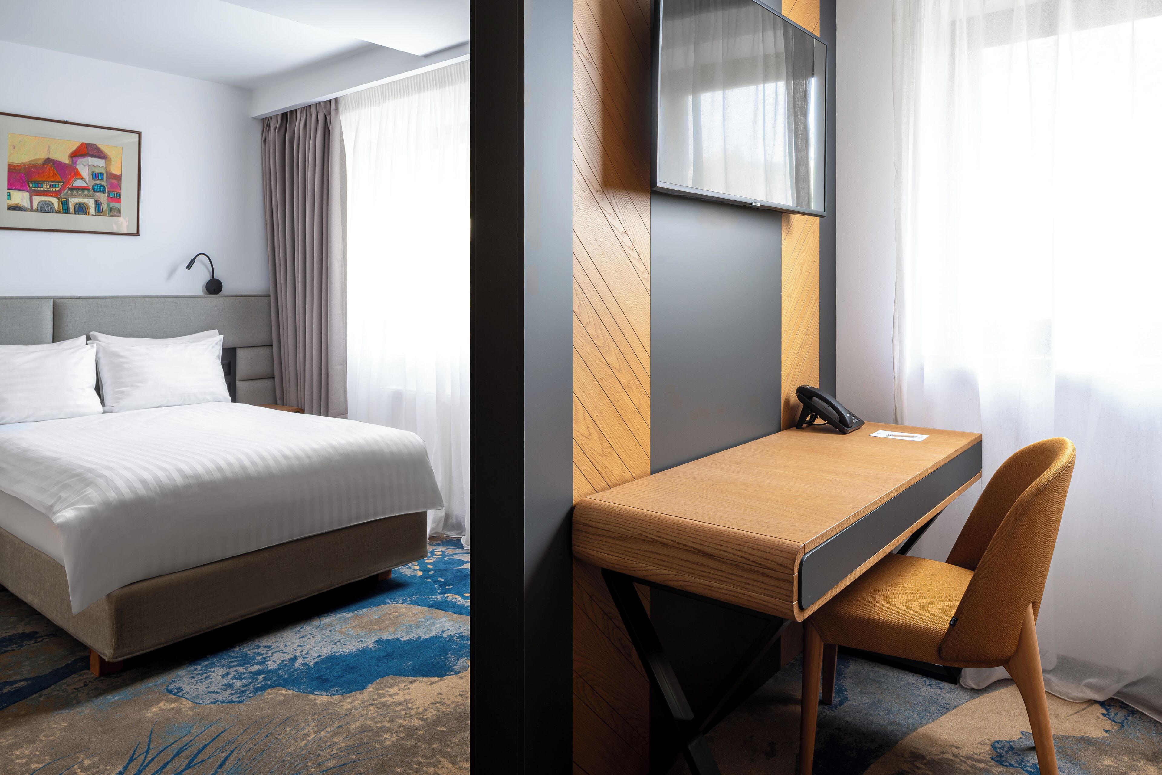 A hotel room with a bed, a desk with a chair, a TV, and a painting on the wall.