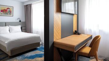 A hotel room with a bed, a desk with a chair, a TV, and a painting on the wall.