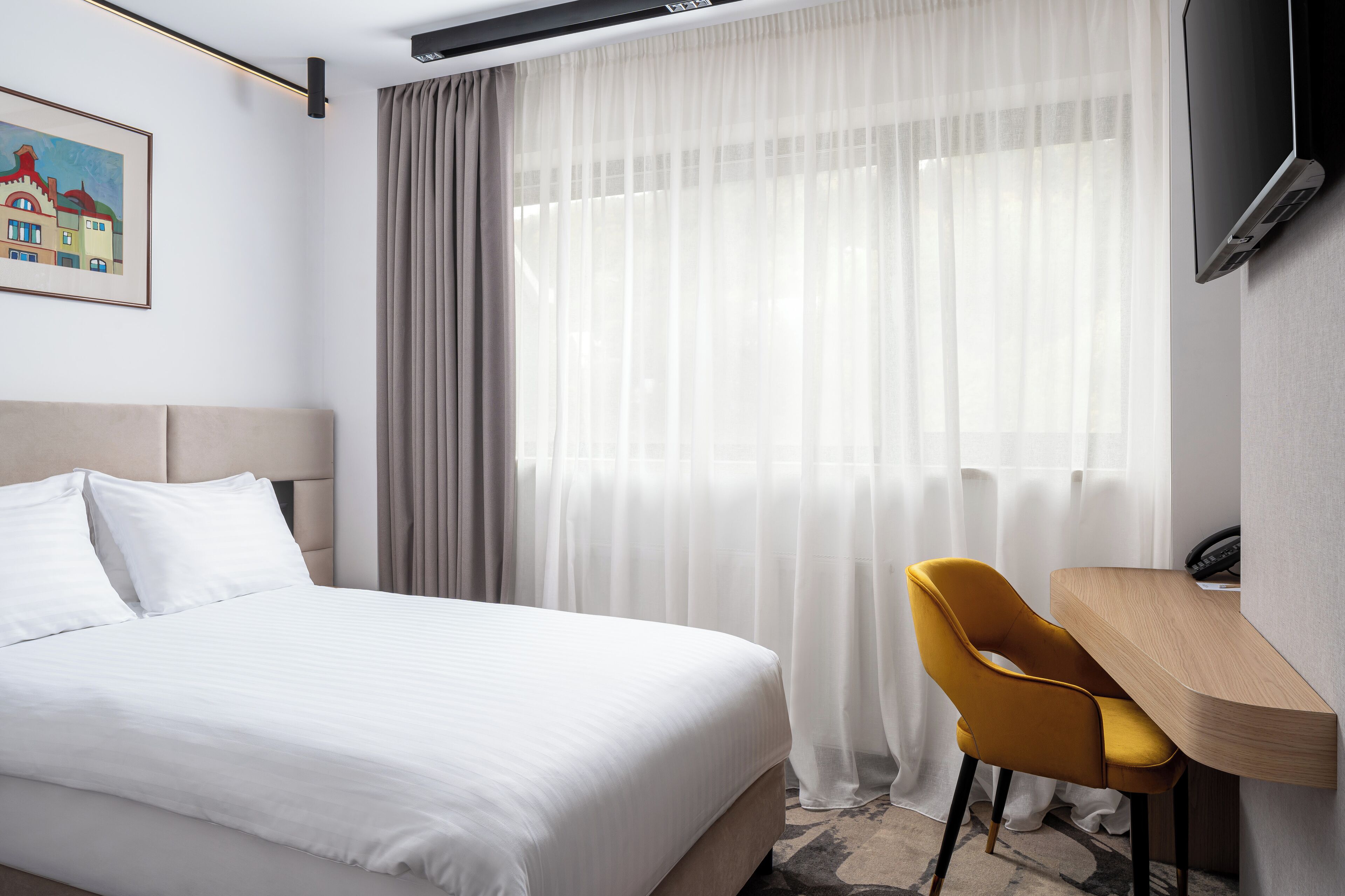 A hotel room with a bed, a desk, a chair, a TV, and a window with sheer curtains.